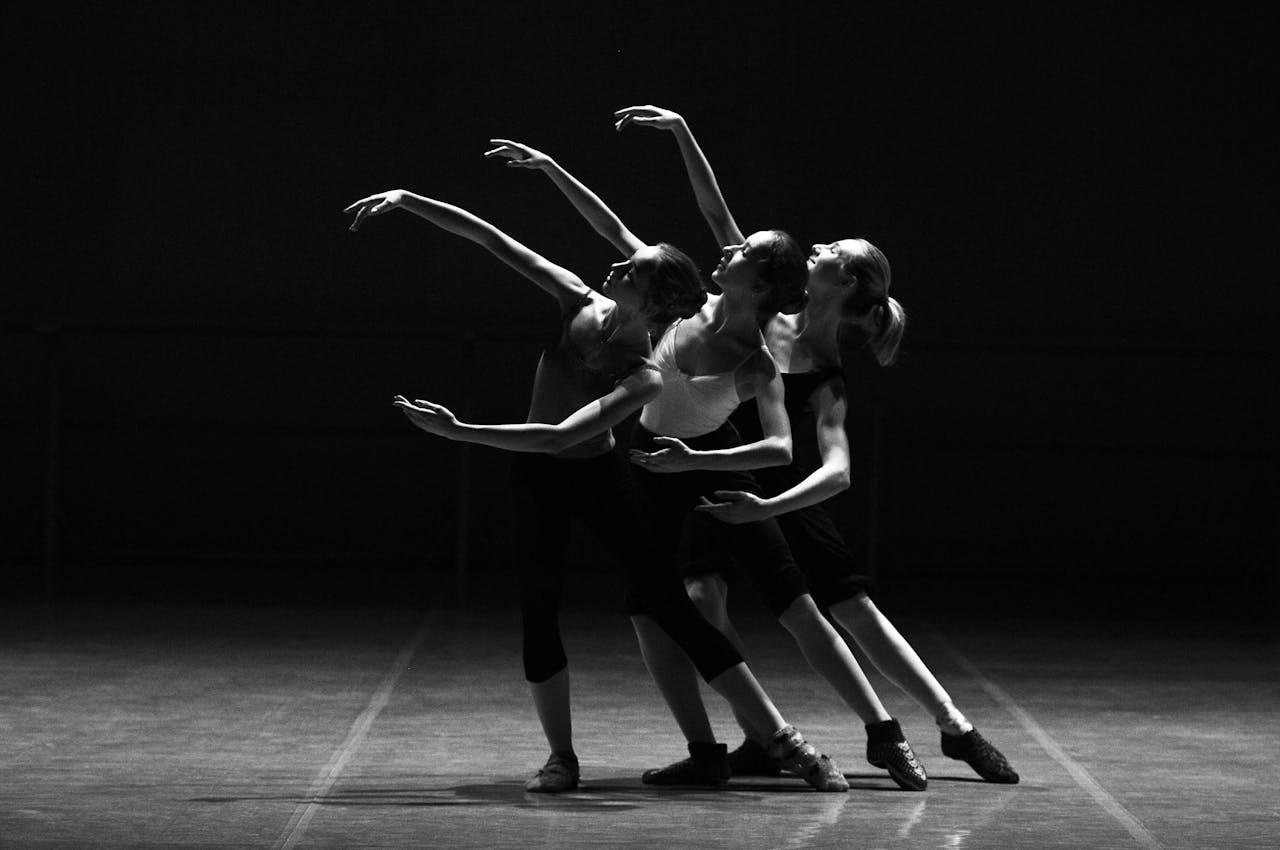 Home Three female ballerinas perform a synchronized dance on stage in dramatic lighting.